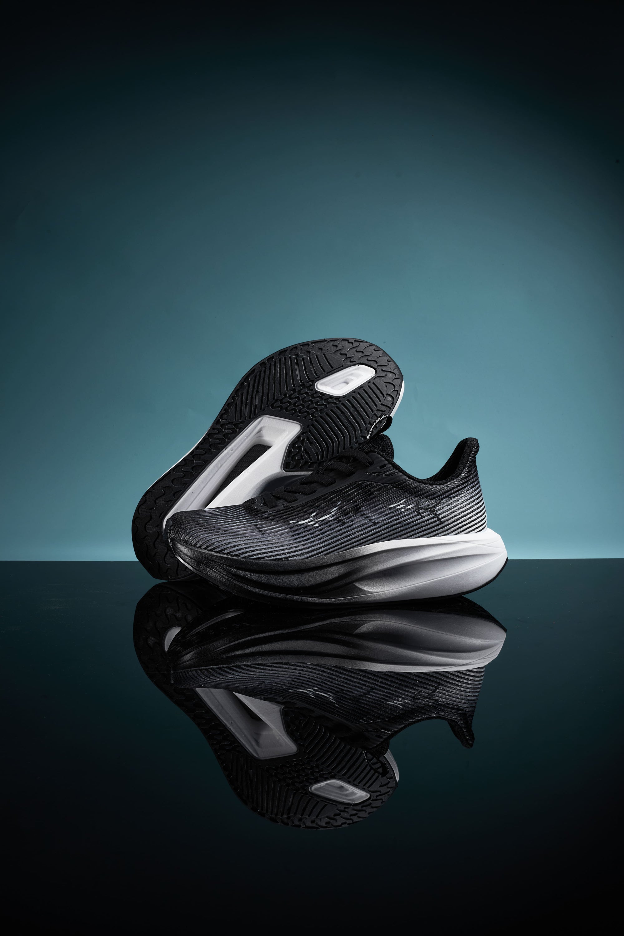 Black sports shoes with a sleek design, featuring a patterned upper and a contrasting white midsole, displayed against a teal background with a reflective surface.
